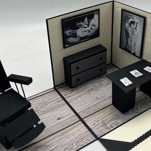 May include: A black and white pop-up card depicting a tattoo parlor with a black leather chair, a black desk with tattoo designs, a black dresser, and two black and white framed pictures of tattoos.