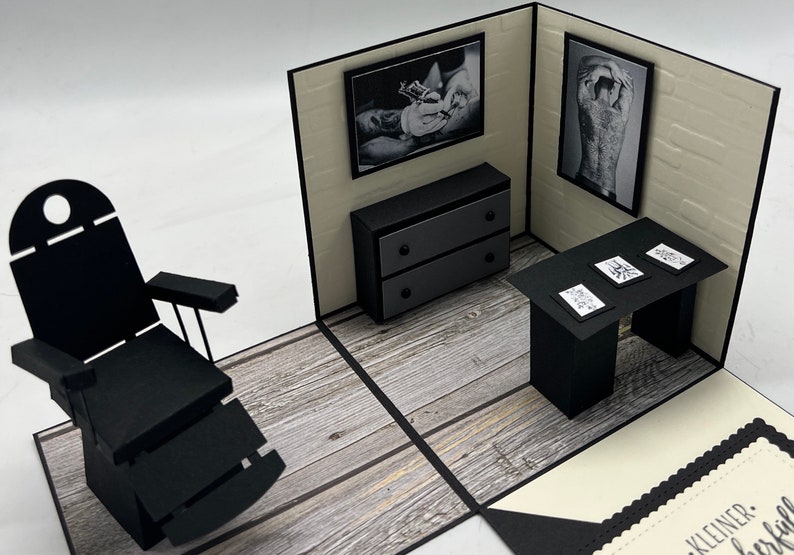 May include: A black and white pop-up card depicting a tattoo parlor with a black leather chair, a black desk with tattoo designs, a black dresser, and two black and white framed pictures of tattoos.