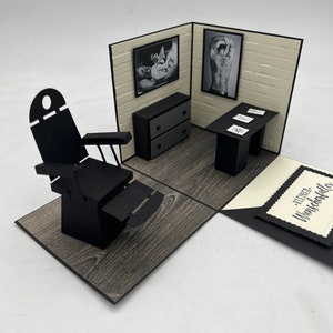 May include: A black and white pop-up card with a 3D model of a tattoo parlor. The parlor features a black leather chair, a black desk with a white brick wall backdrop, and two black and white framed pictures. The card has a black and white tag with the text "Kleiner Wunschk&uuml;nstler".