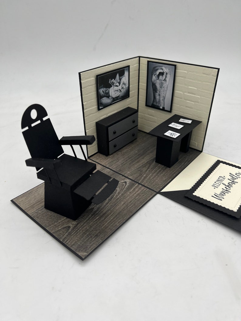 May include: A black and white pop-up card with a 3D model of a tattoo parlor. The parlor features a black leather chair, a black desk with a white brick wall backdrop, and two black and white framed pictures. The card has a black and white tag with the text "Kleiner Wunschk&uuml;nstler".