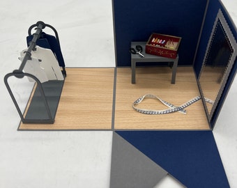 May include: A pop-up card with a tailor shop theme. The card features a small wooden table with a red box of thread, a pair of scissors, and a measuring tape. There is a rack of clothes and a mirror in the background. The card is open and the inside is blank.