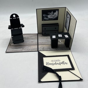 May include: A black and white papercraft pop-up card with a small room interior. The room has a black chair, a dresser with two drawers, a table with three framed pictures, and a small black rug. The card is open and the inside has a white background with a black border and the text "KLEINER WUNSCHERF&Uuml;LLER".