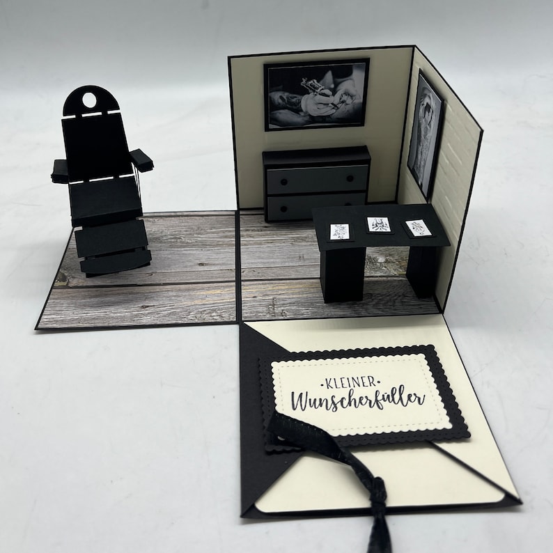 May include: A black and white papercraft pop-up card with a small room interior. The room has a black chair, a dresser with two drawers, a table with three framed pictures, and a small black rug. The card is open and the inside has a white background with a black border and the text "KLEINER WUNSCHERF&Uuml;LLER".