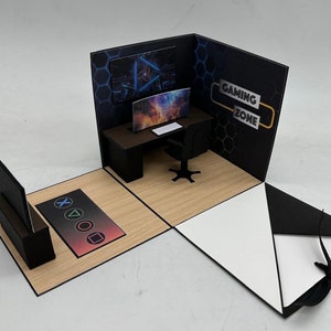 Könnte beinhalten: Eine Pop-up-Karte mit einem Gaming-Thema. Die Karte zeigt eine Schwarzweißillustration eines Gaming-Setups mit einem Computermonitor, einer Tastatur und einem Stuhl. Die Karte enthält auch den Text "Gaming Zone".