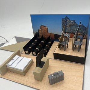 Könnte beinhalten: Ein Miniatur-Papiermodell eines Hotelzimmers mit Bett, Fernseher, Koffer und einer Bühne mit Stühlen und Musikinstrumenten. Der Hintergrund ist ein Foto einer Stadtkulisse.