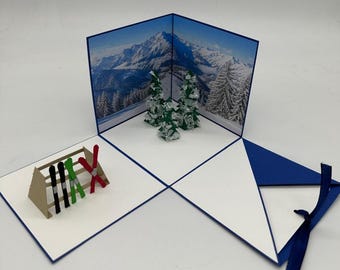 May include: A three-dimensional pop-up greeting card featuring a snowy mountain landscape backdrop, three small green paper pine trees, and a brown paper ski rack holding two black skis, one green ski, and one red pair of crossed skis.