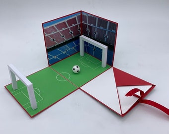 May include: A red and white paper box shaped like a soccer field with a white soccer ball and two white goal posts. The box is open and the inside is white.