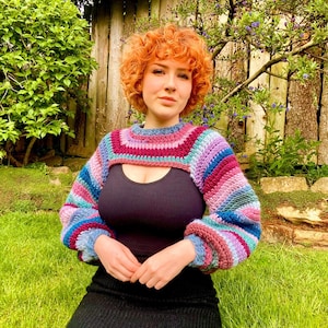 May include: A colorful, crocheted shrug with a cropped design. The sleeves are long and puffy, and the color scheme includes shades of blue, purple, pink, and burgundy. The garment is worn over a black top.