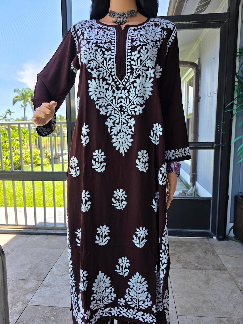 May include: A long, dark brown dress with white floral embroidery. The dress features long sleeves and a round neckline with a V-shaped opening. The floral design is concentrated around the neckline, sleeves, and hem.