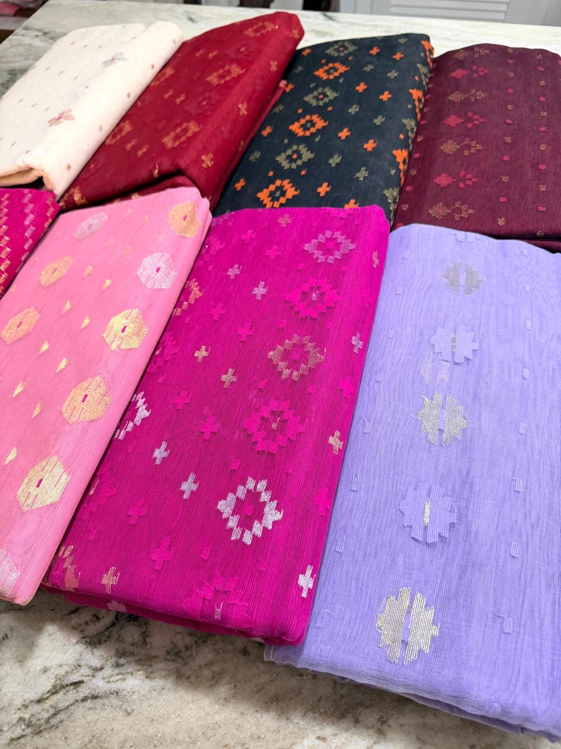 May include: A collection of colorful fabrics, each folded and displaying unique patterns. The fabrics range in color from light pink to deep burgundy, with gold and silver metallic accents. The patterns include geometric shapes and floral designs.
