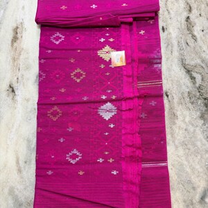 May include: A vibrant fuchsia textile with intricate geometric patterns in gold and silver. The fabric is folded, revealing the detailed design and texture. This textile is likely a traditional garment.