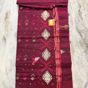 May include: A burgundy textile with intricate gold and red geometric patterns. The fabric is folded, revealing a top section and a patterned main body. The design includes diamond and floral motifs, with a striped border and a tag attached.