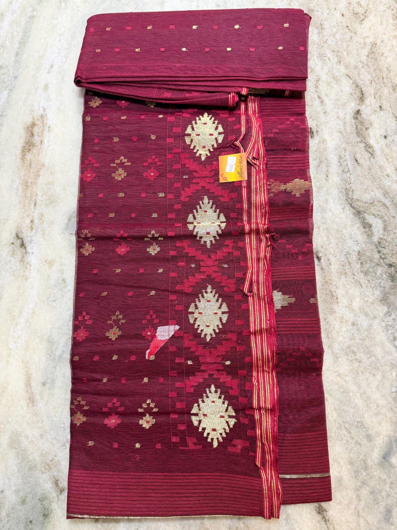 May include: A burgundy textile with intricate gold and red geometric patterns. The fabric is folded, revealing a top section and a patterned main body. The design includes diamond and floral motifs, with a striped border and a tag attached.