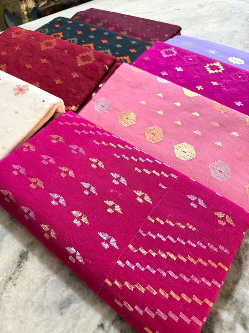 May include: A collection of folded fabric pieces in various colors, including fuchsia, pink, red, and navy. The fabrics feature intricate patterns with metallic accents, such as gold and silver, creating a textured and decorative appearance. The textiles are likely used for clothing or home decor.