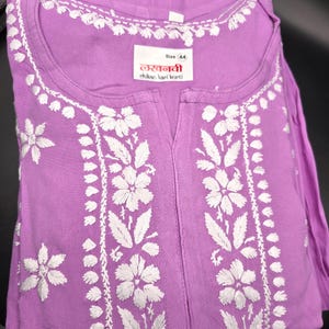 May include: A lavender tunic with intricate white floral embroidery. The tunic features a round neckline and a split front. The label reads "Size 44" and includes text in a foreign language. The garment is folded and appears to be made of a soft fabric.