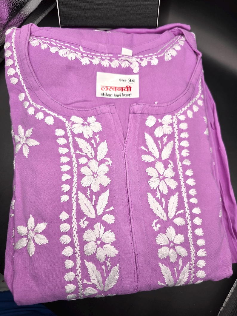 May include: A lavender tunic with intricate white floral embroidery. The tunic features a round neckline and a split front. The label reads "Size 44" and includes text in a foreign language. The garment is folded and appears to be made of a soft fabric.