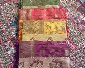 May include: A stack of six colorful silk scarves with intricate patterns and gold accents. The scarves are arranged in a vertical stack, with the bottom scarf being a deep purple color and the top scarf being a light pink color.