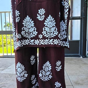 May include: A two-piece maroon outfit with white floral embroidery. The top and pants feature a repeating pattern of stylized trees and flowers. The outfit is made of a soft, flowing fabric, suitable for warm weather.