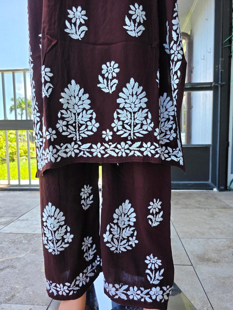 May include: A two-piece maroon outfit with white floral embroidery. The top and pants feature a repeating pattern of stylized trees and flowers. The outfit is made of a soft, flowing fabric, suitable for warm weather.