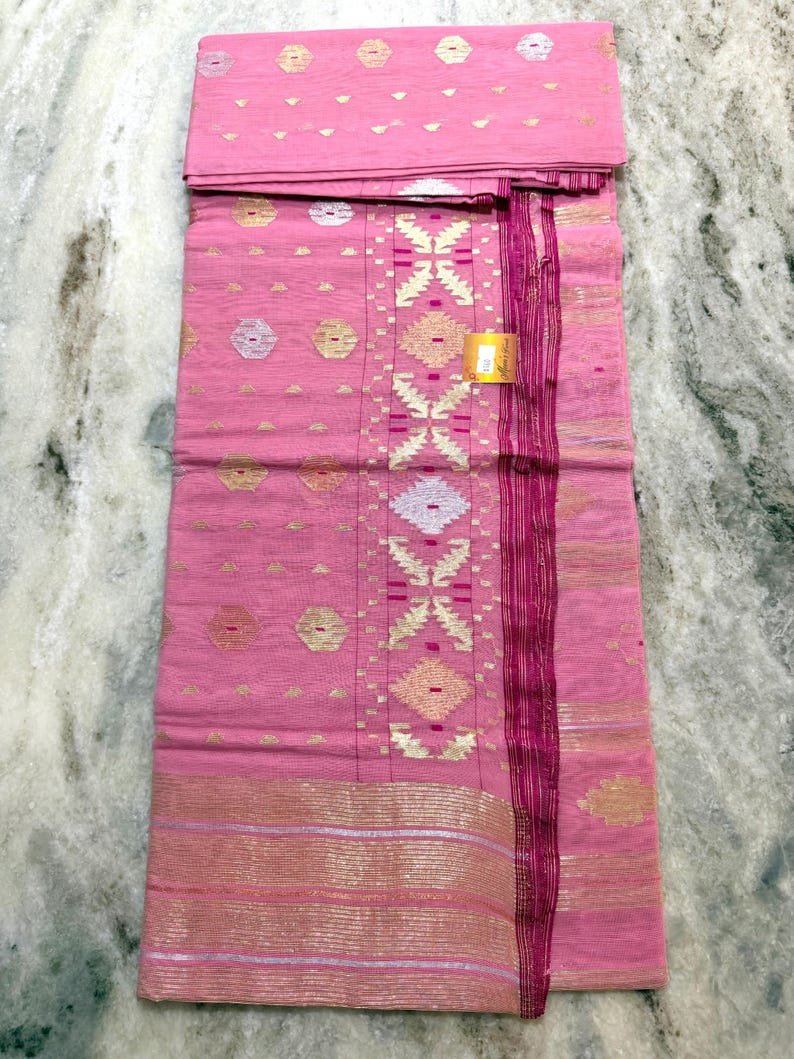 May include: Pink textile with gold and silver geometric patterns. The fabric has a central design of diamond shapes and a border with horizontal stripes. The textile is folded and appears to be new.