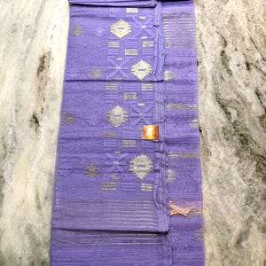 May include: A folded, lavender-colored textile with intricate silver and white geometric patterns. The fabric has a delicate, sheer quality, with a tasseled edge and a small tag attached. The design includes horizontal stripes and floral motifs.