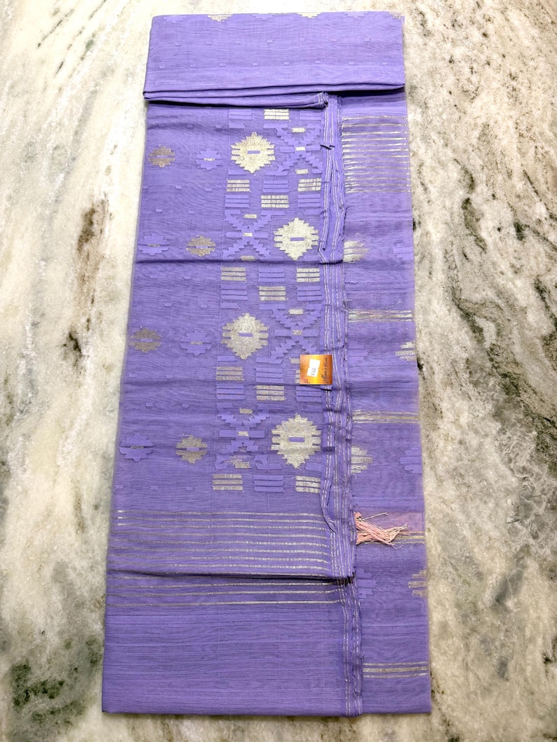 May include: A folded, lavender-colored textile with intricate silver and white geometric patterns. The fabric has a delicate, sheer quality, with a tasseled edge and a small tag attached. The design includes horizontal stripes and floral motifs.