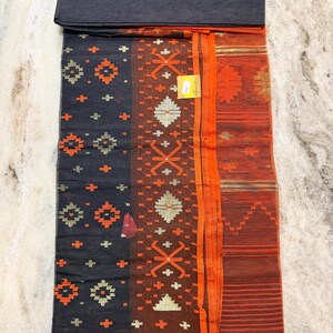May include: A dark gray textile with orange and gold geometric patterns. The fabric features a folded section at the top and a border of orange stripes. The design includes cross and diamond shapes. A small tag is visible.