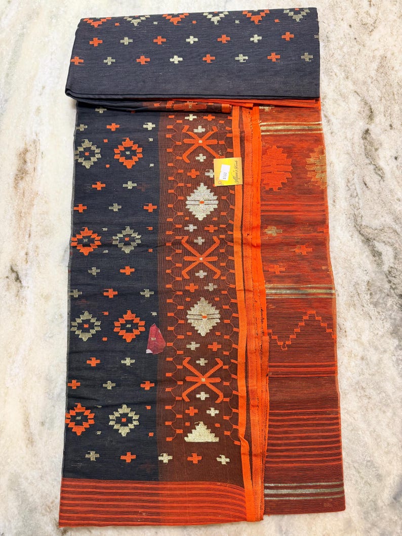 May include: A dark gray textile with orange and gold geometric patterns. The fabric features a folded section at the top and a border of orange stripes. The design includes cross and diamond shapes. A small tag is visible.