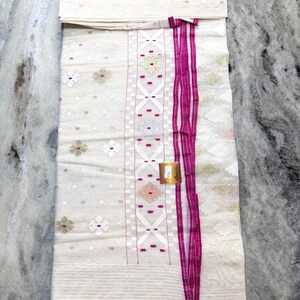 May include: A white fabric with a detailed pattern and a vibrant pink border and accents. The fabric features floral and geometric designs, with a folded section at the top. The overall aesthetic is elegant and detailed.