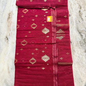 May include: A folded red textile with gold geometric patterns and accents. The fabric features a repeating design of gold shapes and cross-like details. The textile has a striped border with gold lines. A small tag is attached.