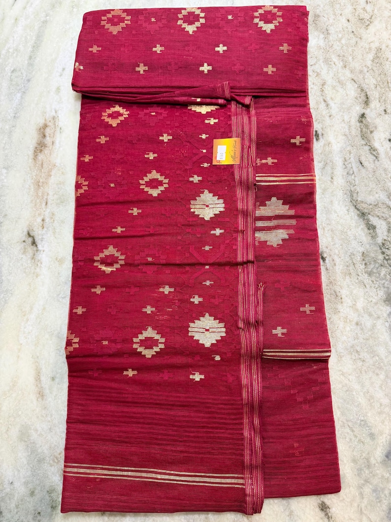 May include: A folded red textile with gold geometric patterns and accents. The fabric features a repeating design of gold shapes and cross-like details. The textile has a striped border with gold lines. A small tag is attached.