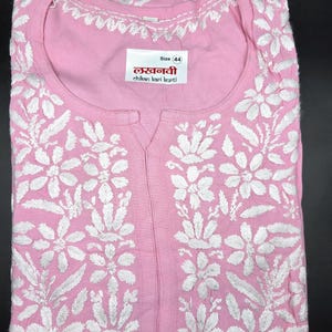 May include: A light pink kurti with intricate white floral embroidery. The kurti features a round neckline and a split front. A label displays the size and text in a foreign language. The fabric is folded neatly.