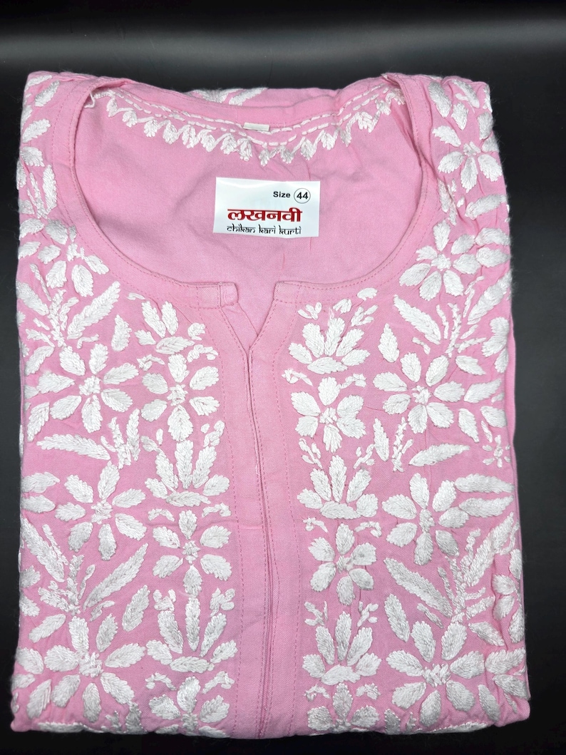 May include: A light pink kurti with intricate white floral embroidery. The kurti features a round neckline and a split front. A label displays the size and text in a foreign language. The fabric is folded neatly.