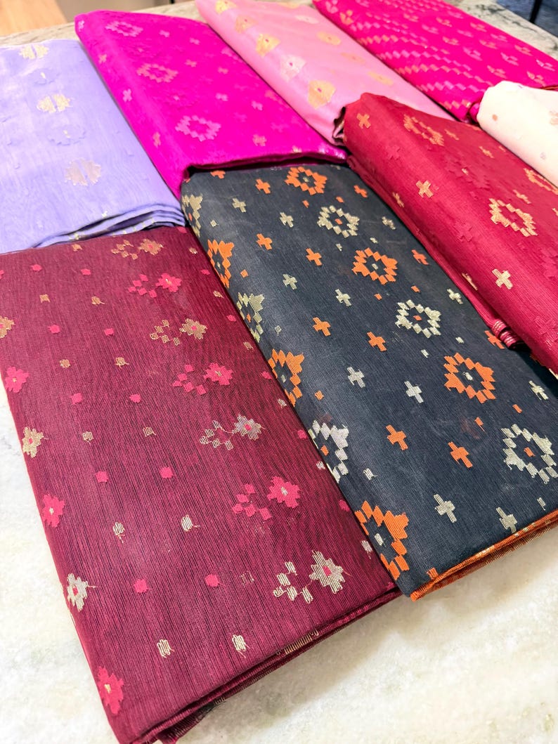 May include: A collection of folded fabric in various colors, including purple, pink, and burgundy. The fabrics feature floral and geometric patterns in contrasting colors. The textiles appear to be lightweight and suitable for clothing or home decor projects.