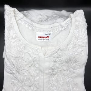 May include: White embroidered tunic with floral and leaf patterns. The tunic has a mandarin collar and a small slit at the neckline. The label reads "Size 42" and includes text in a foreign language. The garment is folded and displayed against a black background.