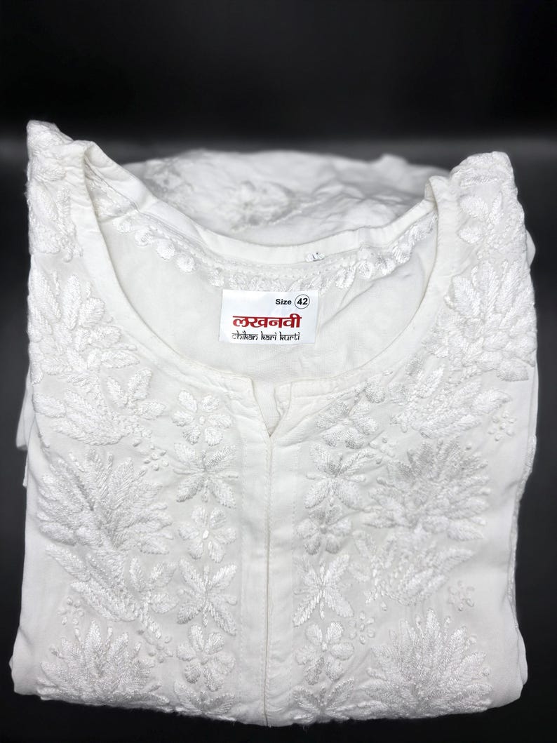 May include: White embroidered tunic with floral and leaf patterns. The tunic has a mandarin collar and a small slit at the neckline. The label reads "Size 42" and includes text in a foreign language. The garment is folded and displayed against a black background.