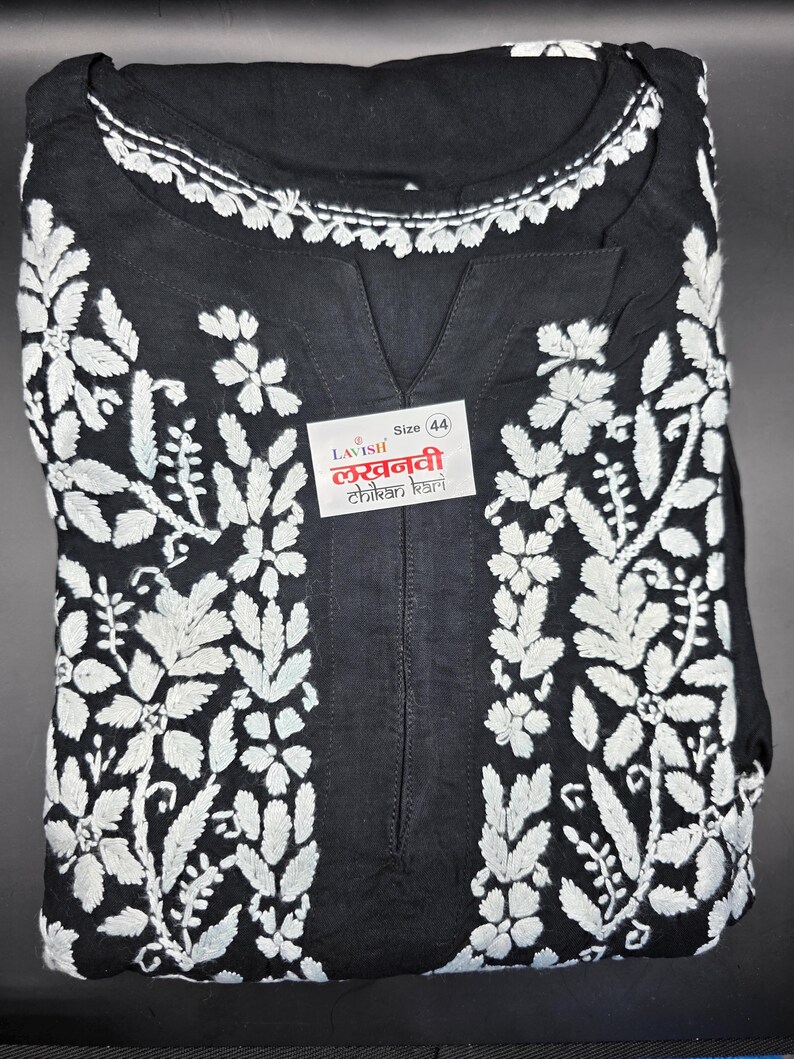 May include: Black tunic with white floral embroidery. The tunic features a V-neck and a white embroidered neckline. A label with text is visible on the front. Size 44.
