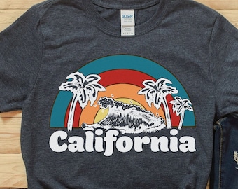 Op de afbeelding: Een donkerblauw T-shirt met een retro-grafische afbeelding van een golf die onder een regenboog met palmbomen breekt, en de tekst "California".