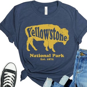 May include: A navy blue t-shirt with a yellow silhouette of a bison and the text "Yellowstone National Park Est. 1872". The shirt is paired with blue jeans and black sneakers.