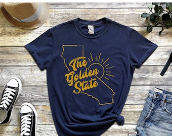 Op de afbeelding: Een marineblauw t-shirt met een gouden omtrek van de staat Californië en de tekst "The Golden State" in een gouden, verweerd lettertype. De omtrek van Californië is gedeeltelijk gevuld met een gouden zon.