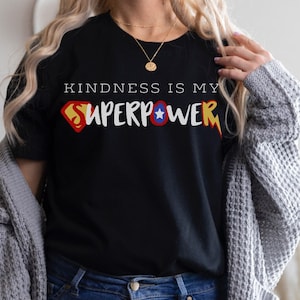 May include: Black t-shirt with the text "KINDNESS IS MY SUPERPOWER" in white, yellow, red, and blue. The design incorporates a superhero logo. The shirt is paired with blue jeans and a gray cardigan.