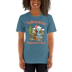 May include: A teal blue t-shirt with a vintage-style graphic of a mountain scene with a waterfall, a pine tree, and a red sun. The text "Yellowstone National Park" is printed in a peachy-orange color.