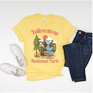 May include: Yellow t-shirt with a vintage-style illustration of a mountain scene with a waterfall, a bear, and a pine tree. The text "Yellowstone National Park" is printed in red and white on the shirt.