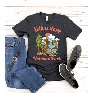 May include: A dark heather grey t-shirt with a vintage-style graphic of a mountain scene with a waterfall, a pine tree, and a red sun. The text "Yellowstone National Park" is printed in orange on the shirt.