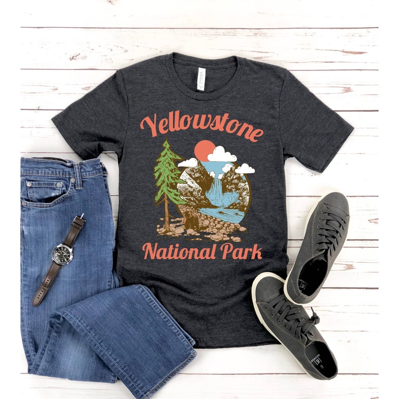 May include: A dark heather grey t-shirt with a vintage-style graphic of a mountain scene with a waterfall, a pine tree, and a red sun. The text "Yellowstone National Park" is printed in orange on the shirt.