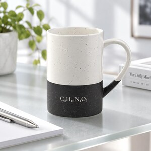 May include: A two-toned ceramic mug featuring a speckled white upper section and a solid black lower section. The black portion displays the white chemical formula C8H10N4O2. The mug includes a matching black handle and a cylindrical body shape.