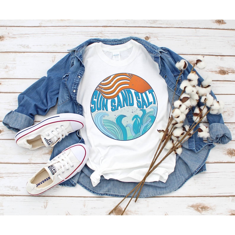 May include: White t-shirt with a blue and orange graphic of waves and the text "Sun Sand Salt". The shirt is laid out on a wooden surface with a denim jacket and white sneakers.