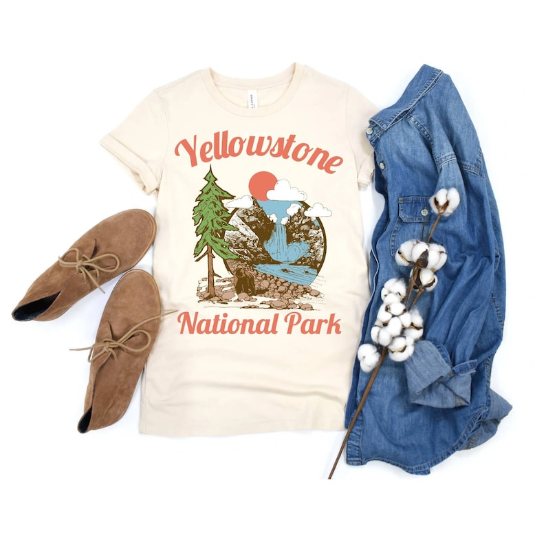 May include: A beige t-shirt with a vintage-style graphic of a mountain scene with a waterfall and a red sun. The text "Yellowstone National Park" is printed in red below the graphic.