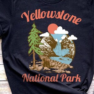 May include: A dark blue t-shirt with a vintage-style illustration of a mountain scene with a waterfall, a pine tree, and a bear. The text "Yellowstone National Park" is printed in red on the shirt.
