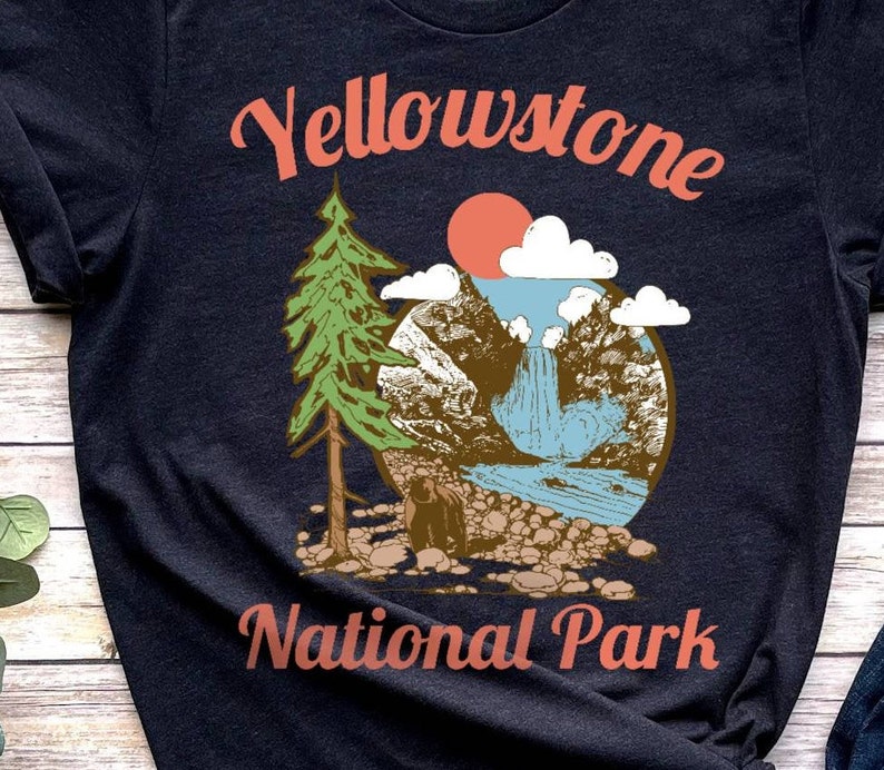 May include: A dark blue t-shirt with a vintage-style illustration of a mountain scene with a waterfall, a pine tree, and a bear. The text "Yellowstone National Park" is printed in red on the shirt.